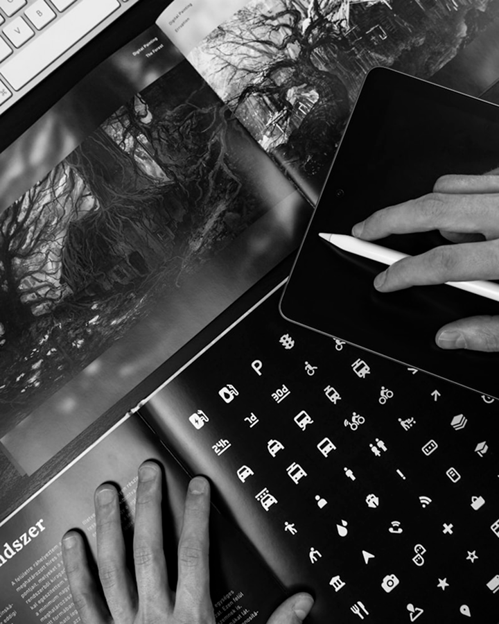 Hand using a stylus on a tablet beside a keyboard and open magazine
